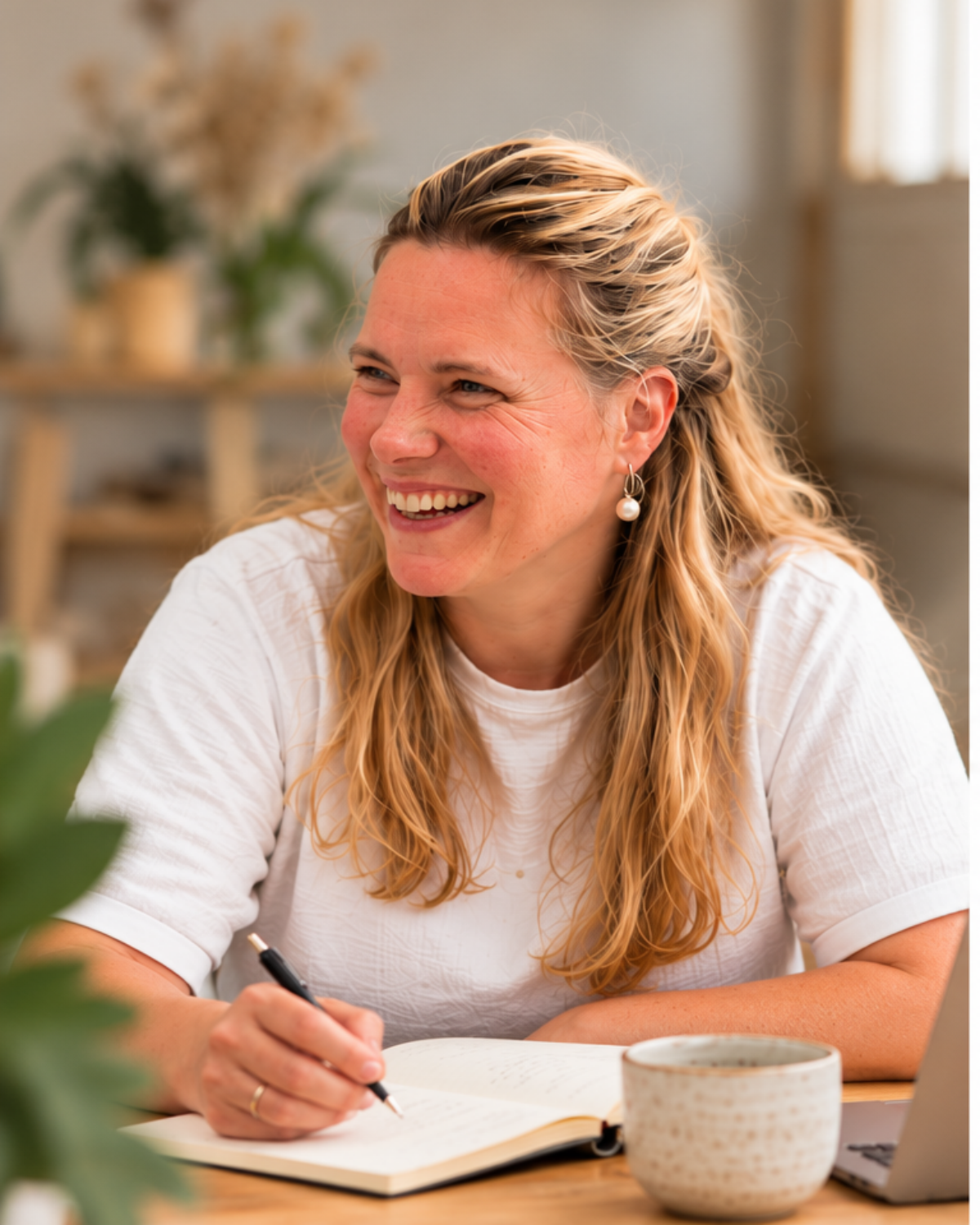 Lian de Snoo zit aan een tafel met een open notitieboek en een pen, en lacht.